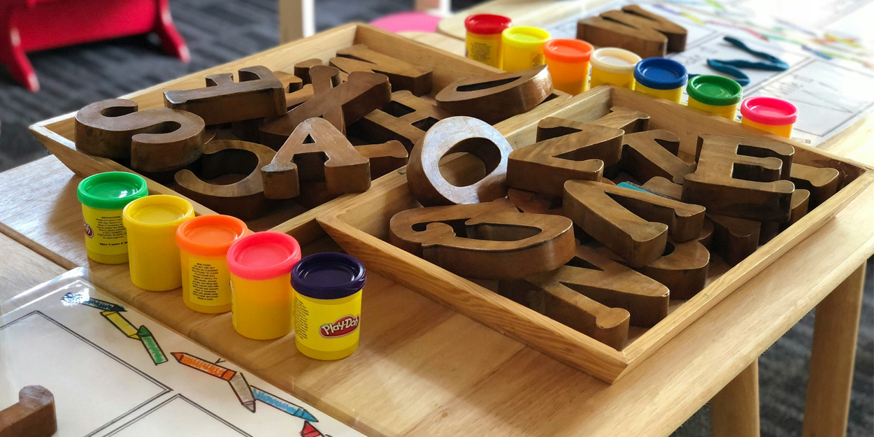 A table set up for a children's activity with trays of brown wooden alphabet letters and Play-Doh cups of different colors